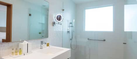 A modern bathroom with a large mirror, a sink with toiletries, a glass shower enclosure, and a window providing natural light.