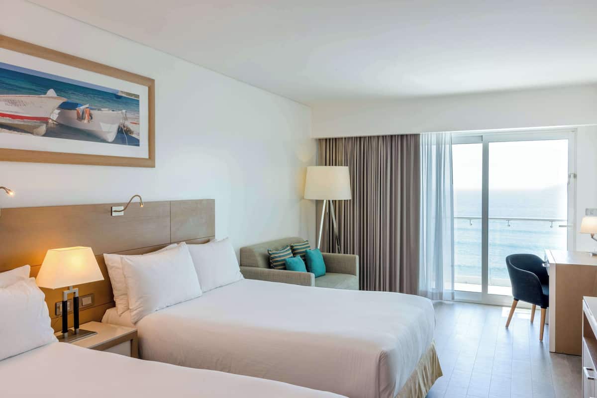 Habitación de hotel con dos camas, un escritorio, una silla, un sofá, una lámpara y un ventanal con vista al mar.