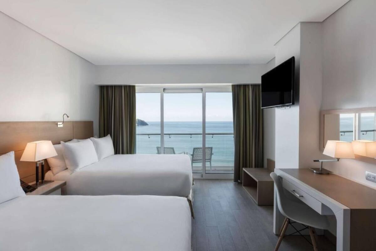 Habitación de hotel con dos camas, un escritorio, televisor y balcón con vista al mar.