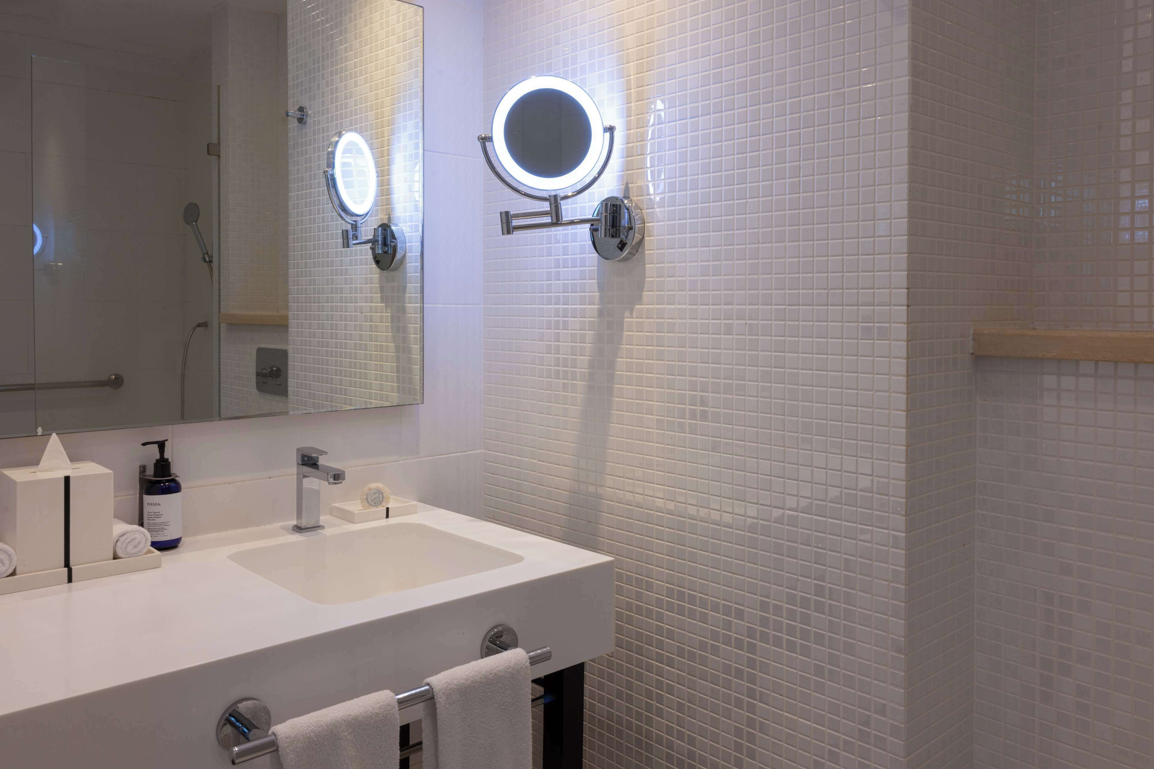 Un baño con lavamanos blanco, dos espejos y un cabezal de ducha manual.