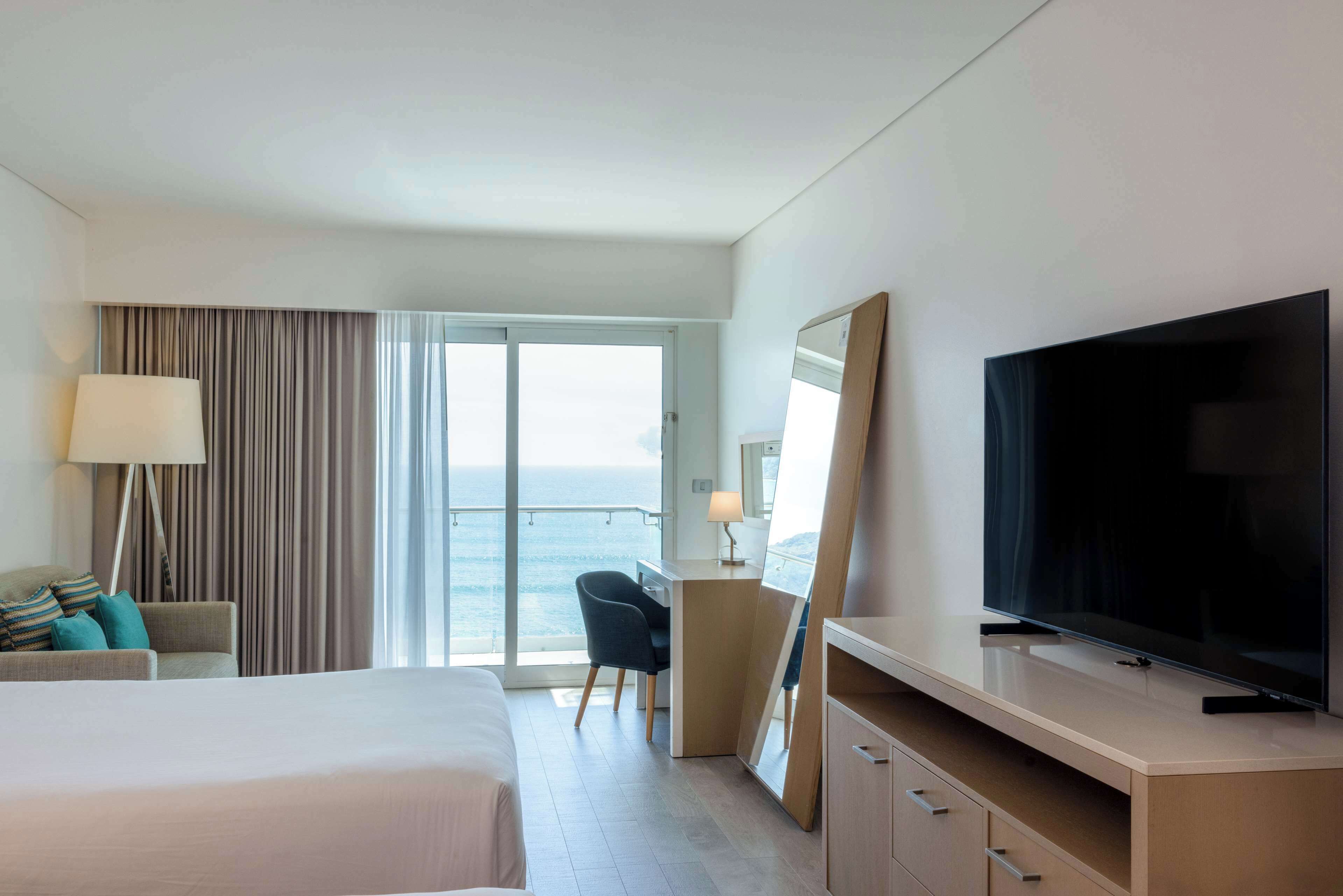 A hotel room with a bed, a desk, a television, a chair, a lamp, and a view of the ocean.