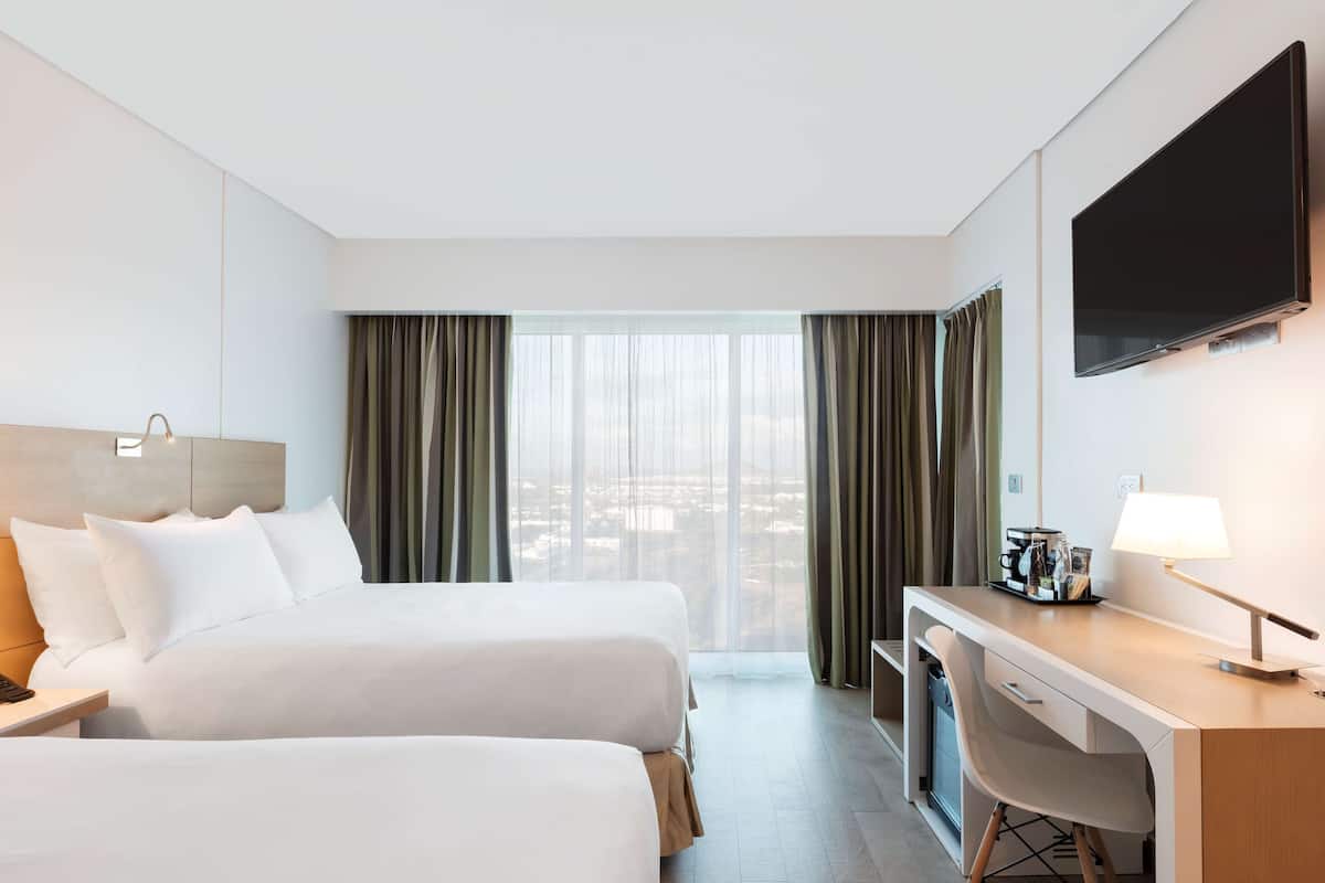 Habitación de hotel con dos camas, un escritorio, televisor y vistas a la ciudad.