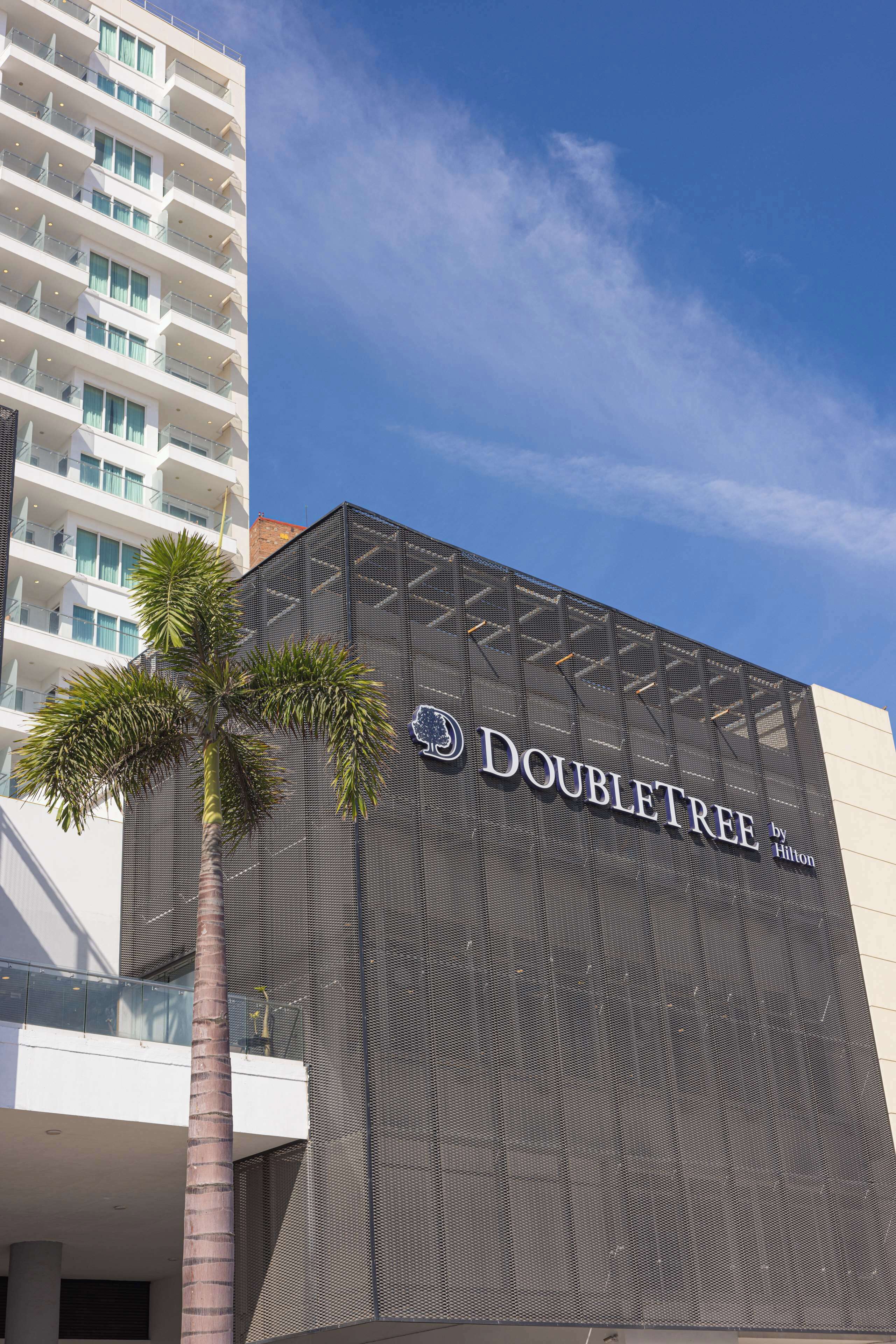 A modern hotel facade with a prominent "DOUBLETREE by Hilton" sign, a palm tree, and a clear blue sky.