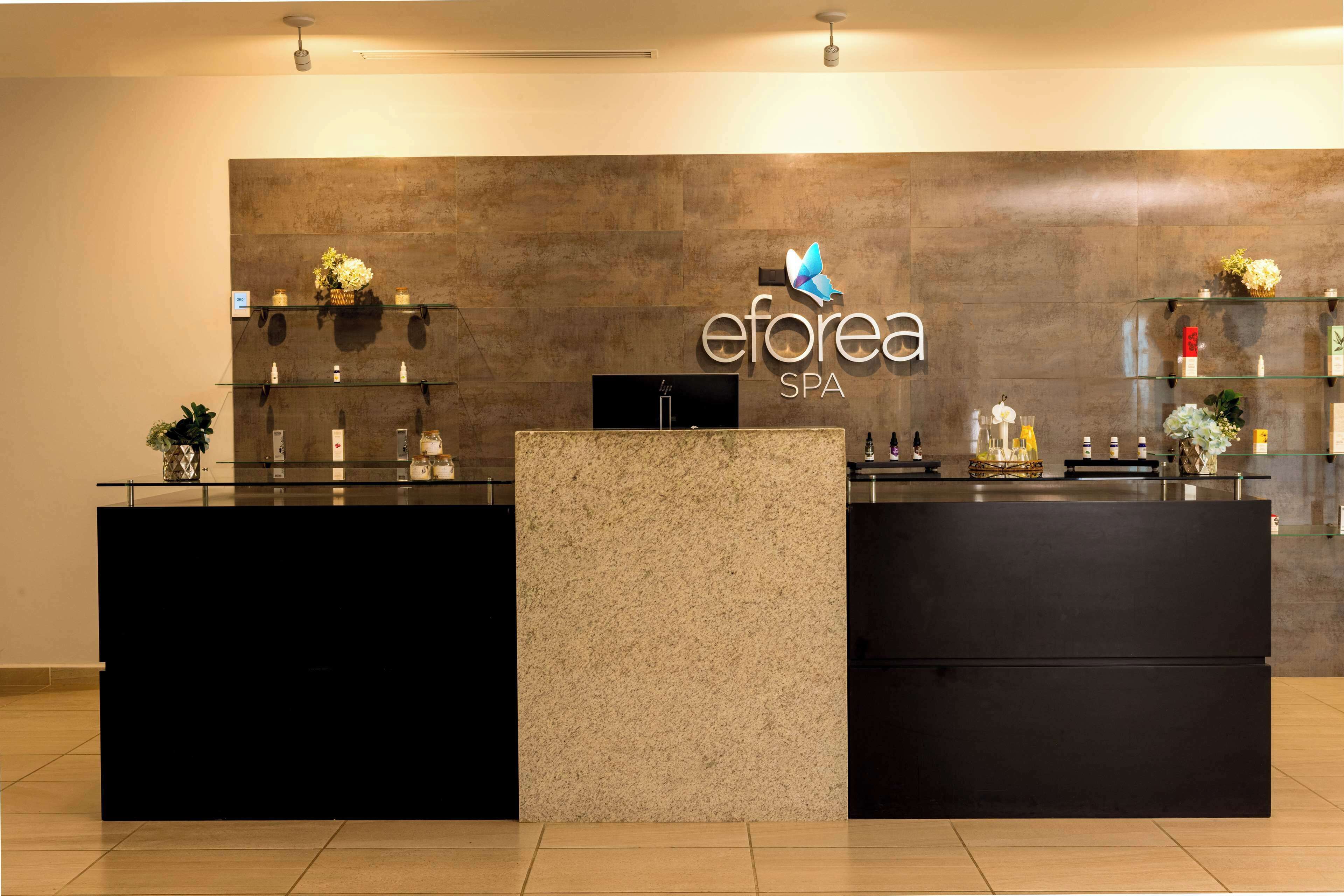 Ein Empfangsschalter mit dem Logo von 'eforea SPA' und einem Schmetterlingssymbol. Auf der linken Seite befinden sich Regale mit verschiedenen Artikeln und eine Pflanze.
