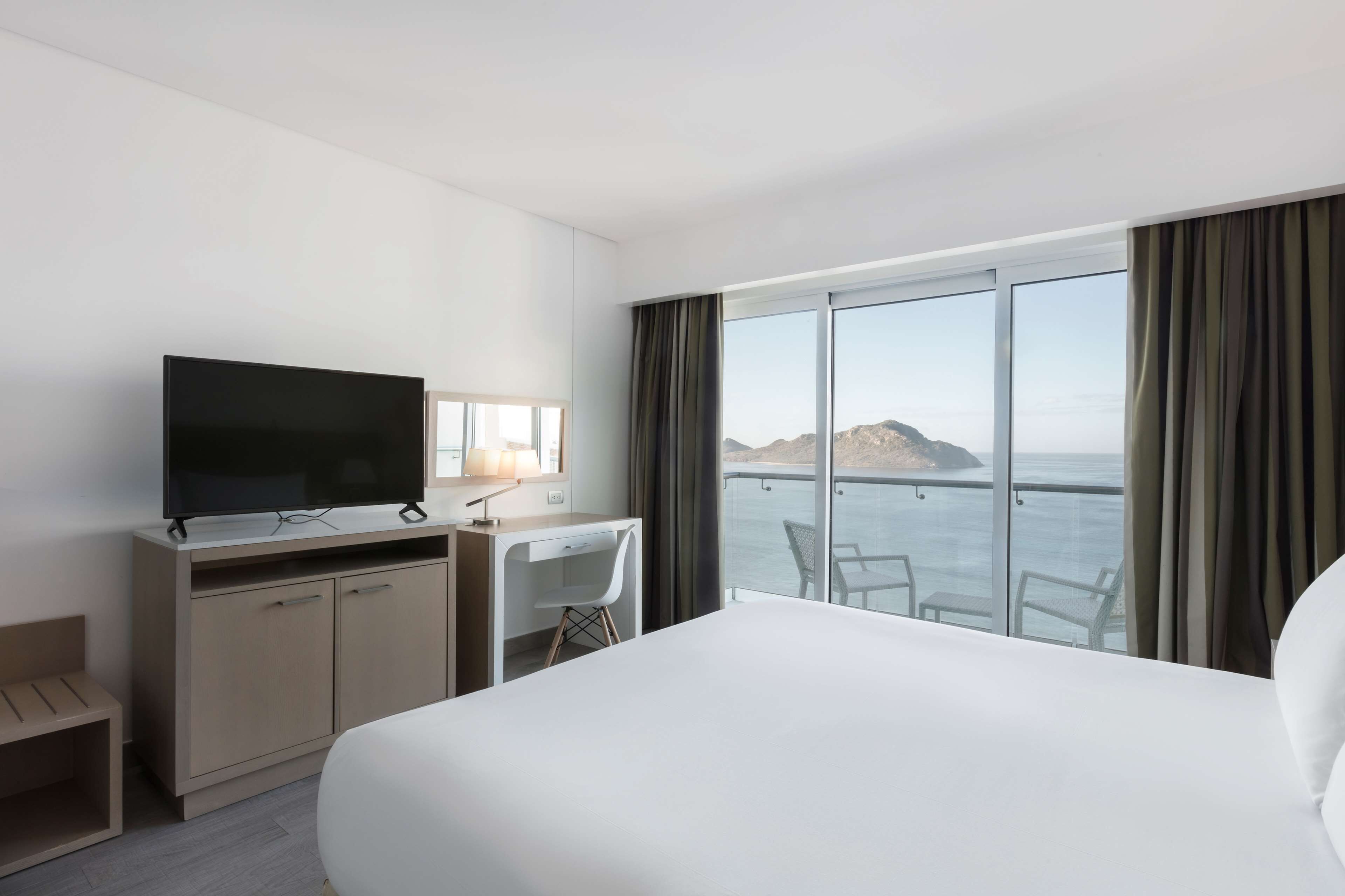 A hotel room with a bed, a desk, a TV, and a large window with a view of the sea.