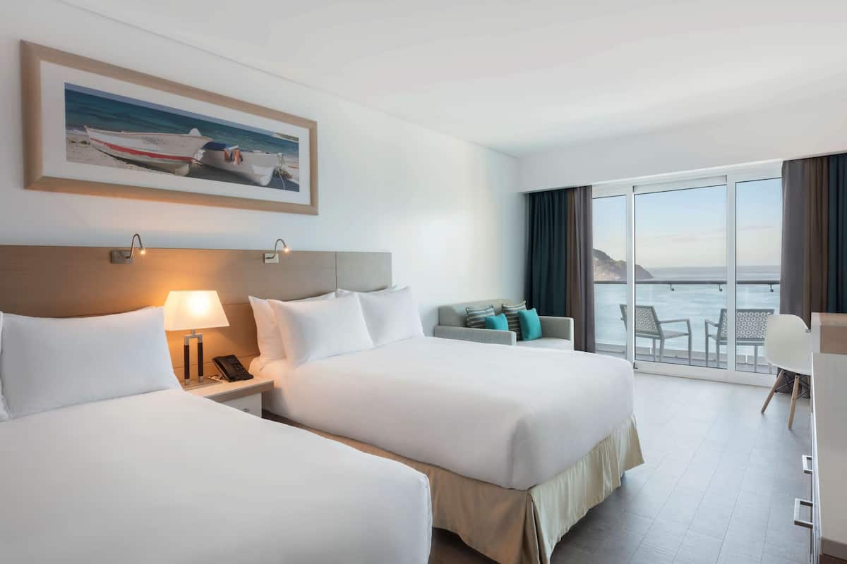 Habitación de hotel con dos camas, una mesita de noche con lámpara, un cuadro enmarcado con una imagen de un barco en la pared, y un balcón con vistas al mar.