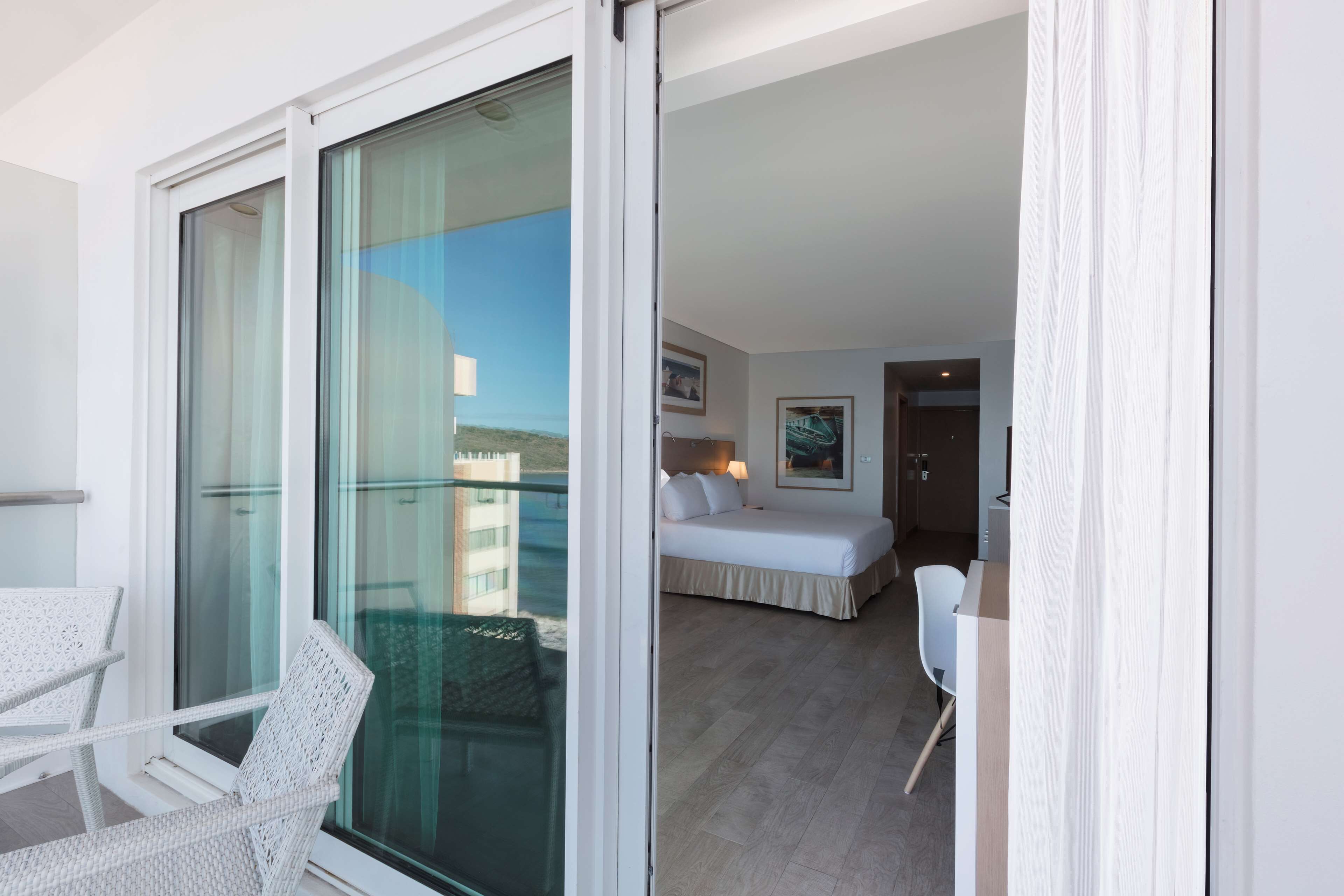 A hotel room with a balcony, a bed, a desk, and a chair.
