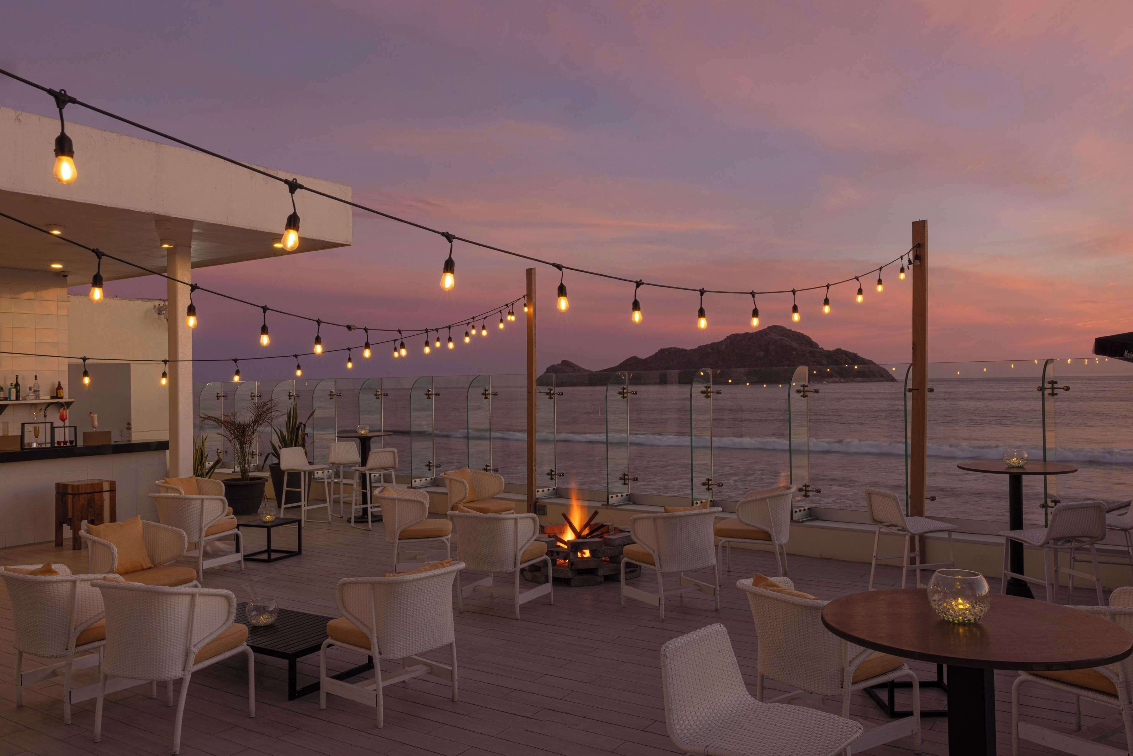 An outdoor seating area with wicker chairs and tables, a fire pit, and string lights against a sunset backdrop.