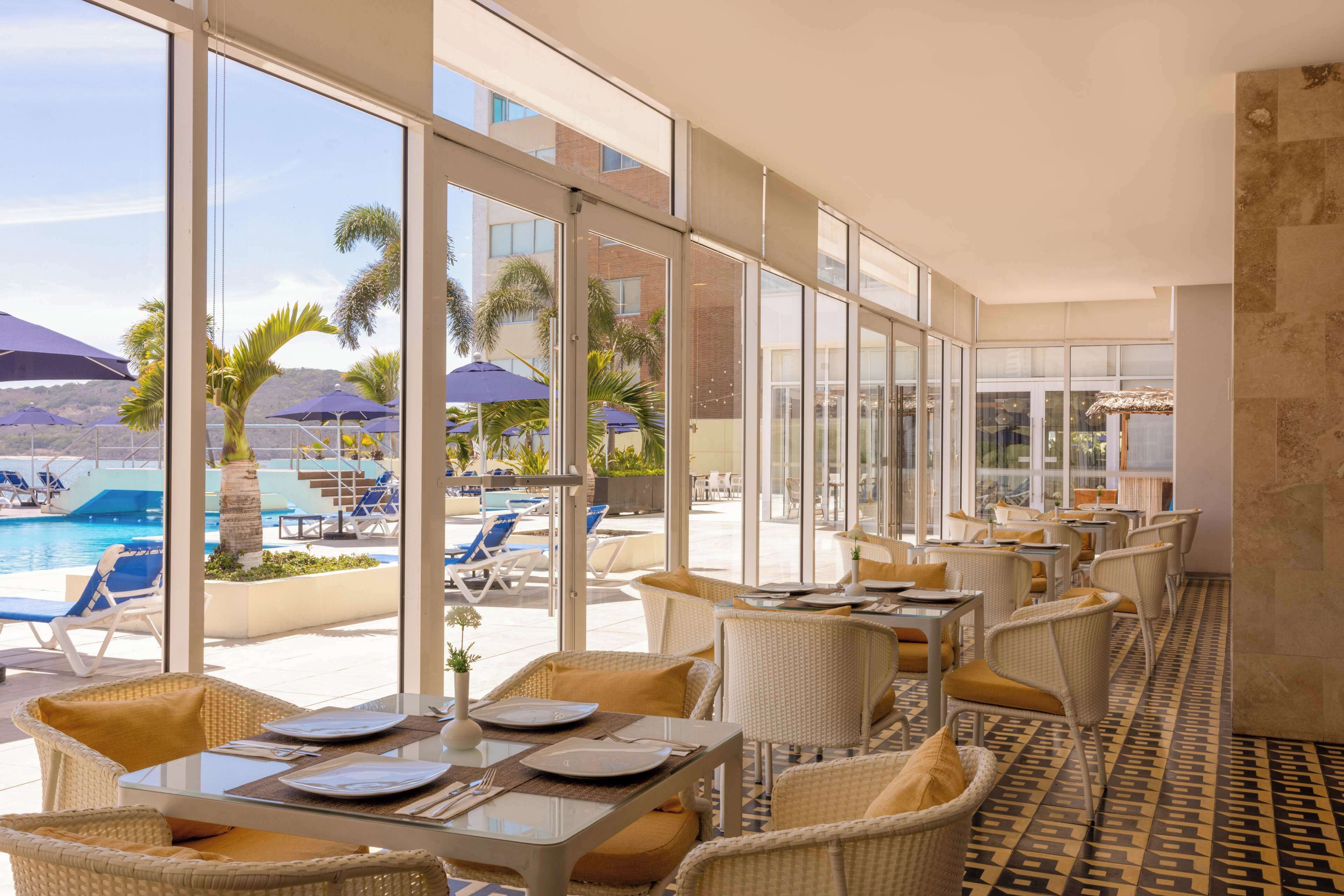 A sunlit outdoor dining area with wicker furniture and large windows overlooking a pool and palm trees.