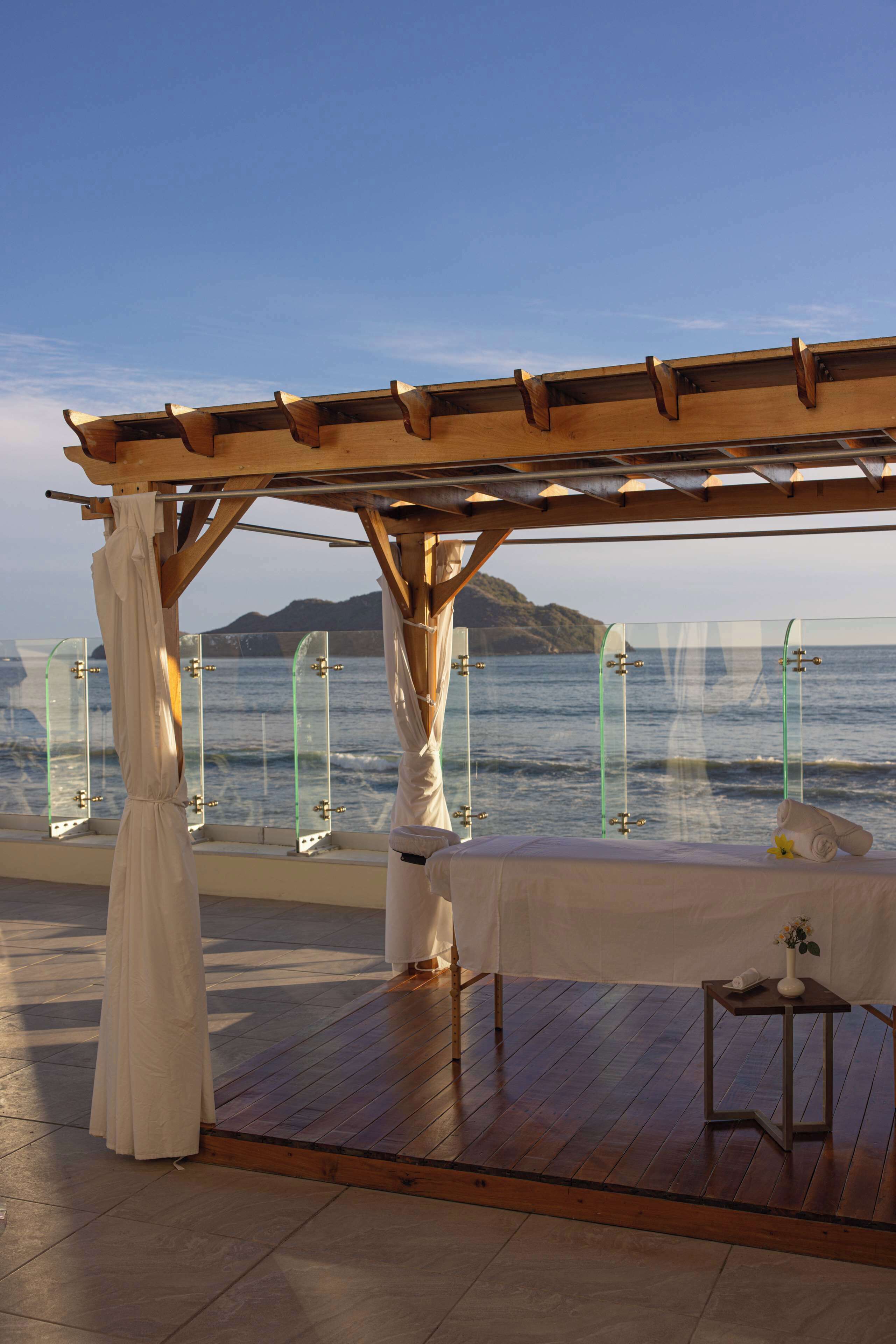 A wooden deck with a pergola, a white table with a vase, and a view of the sea.