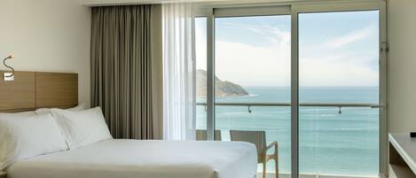 A hotel room with two beds, a large sliding glass door leading to a balcony with a view of the sea and a distant island.