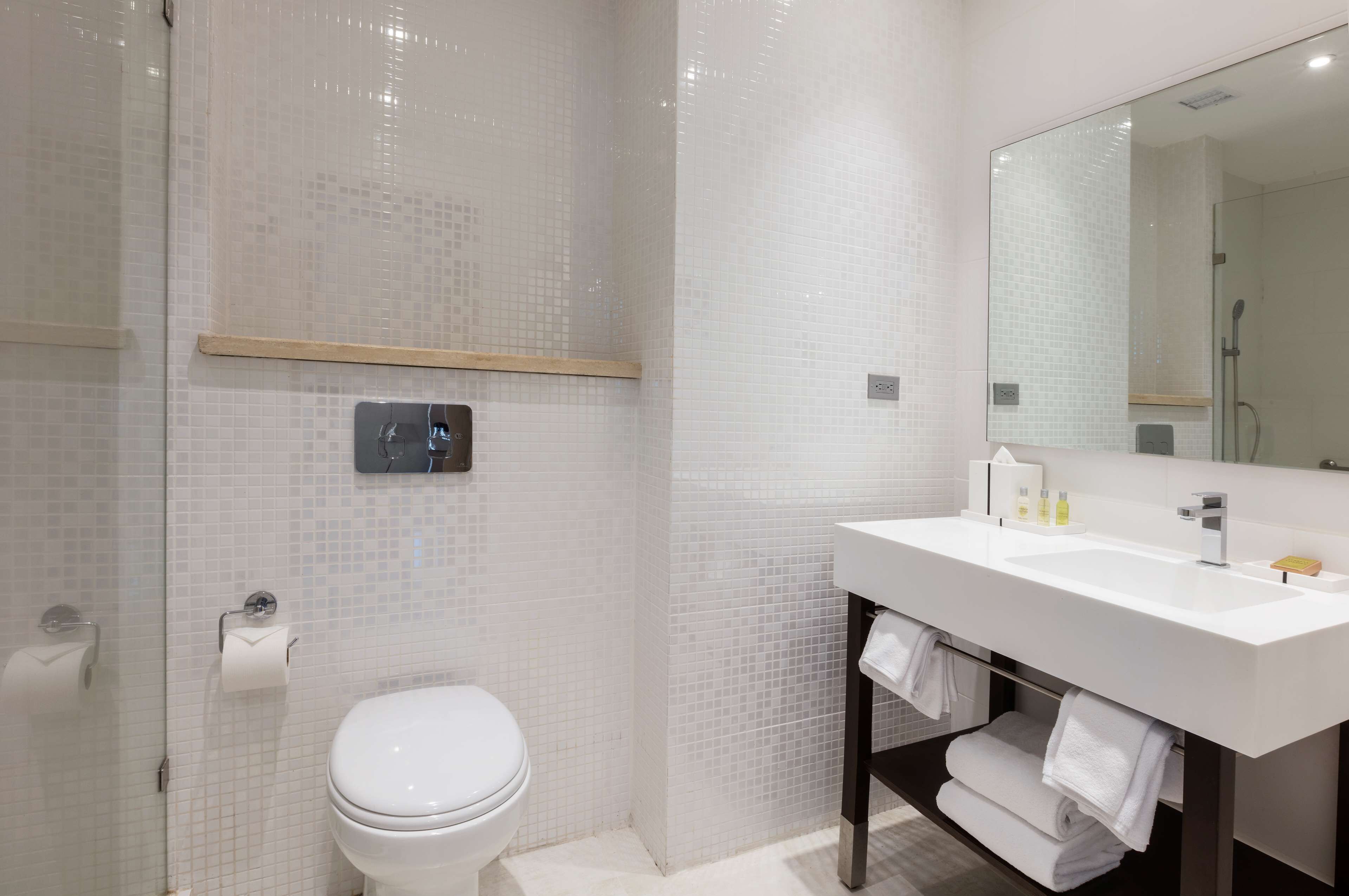 Un baño moderno con ducha de azulejos blancos, inodoro blanco y un espejo grande sobre un lavabo blanco con toallero.