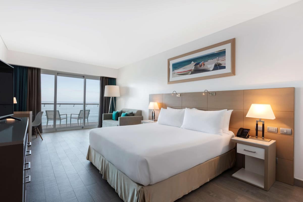 Habitación de hotel con una cama grande, un escritorio y una silla. Tiene un balcón con vista al mar.