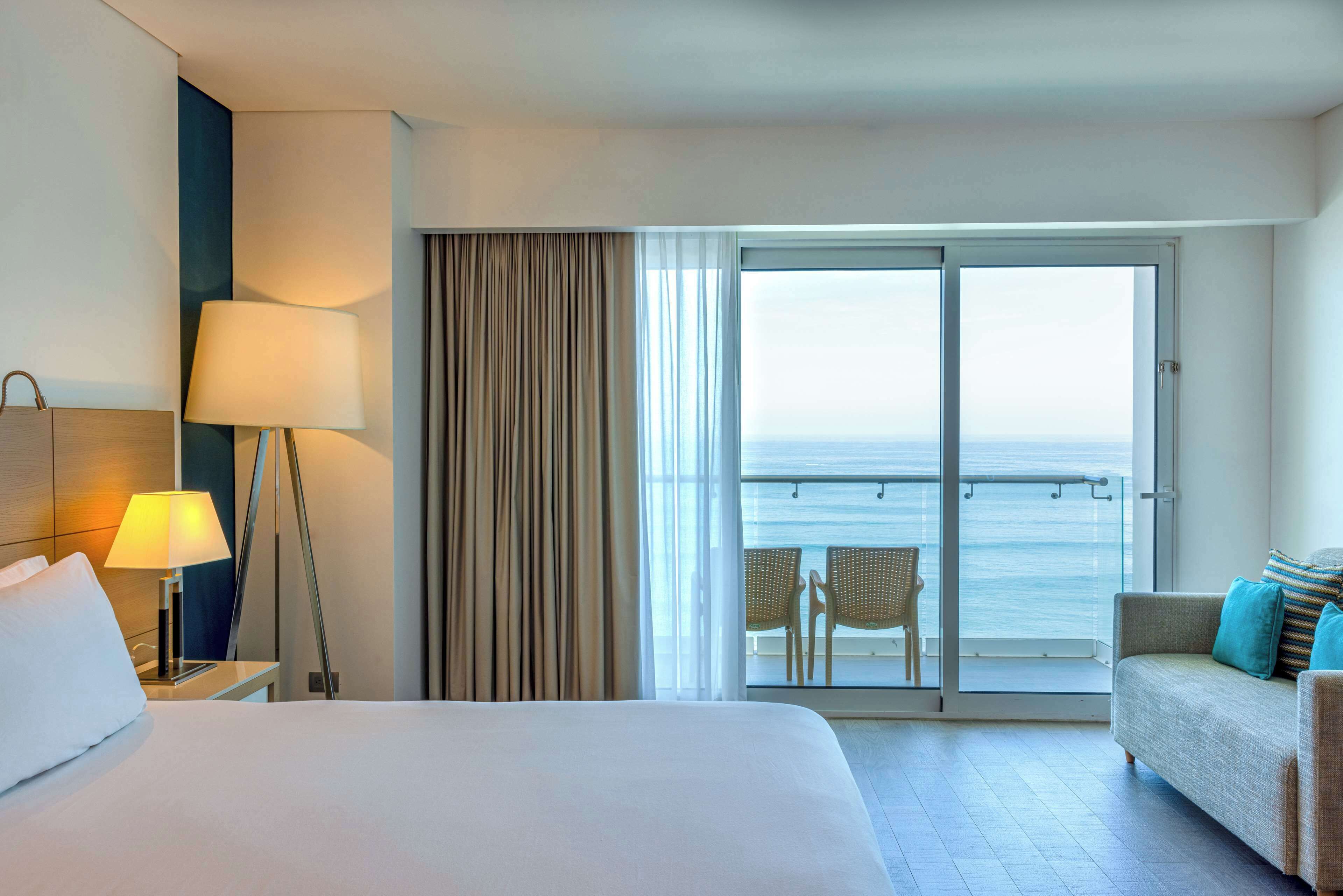 A hotel room with a bed, a nightstand with a lamp, a sofa, and a view of the ocean through a sliding glass door.