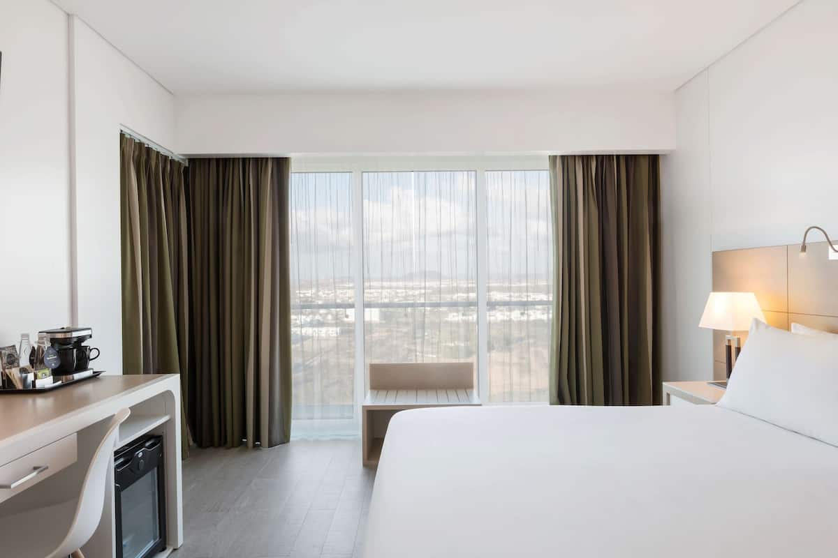 Habitación de hotel con cama, escritorio, silla, lámpara y vista de la ciudad a través de amplias ventanas.