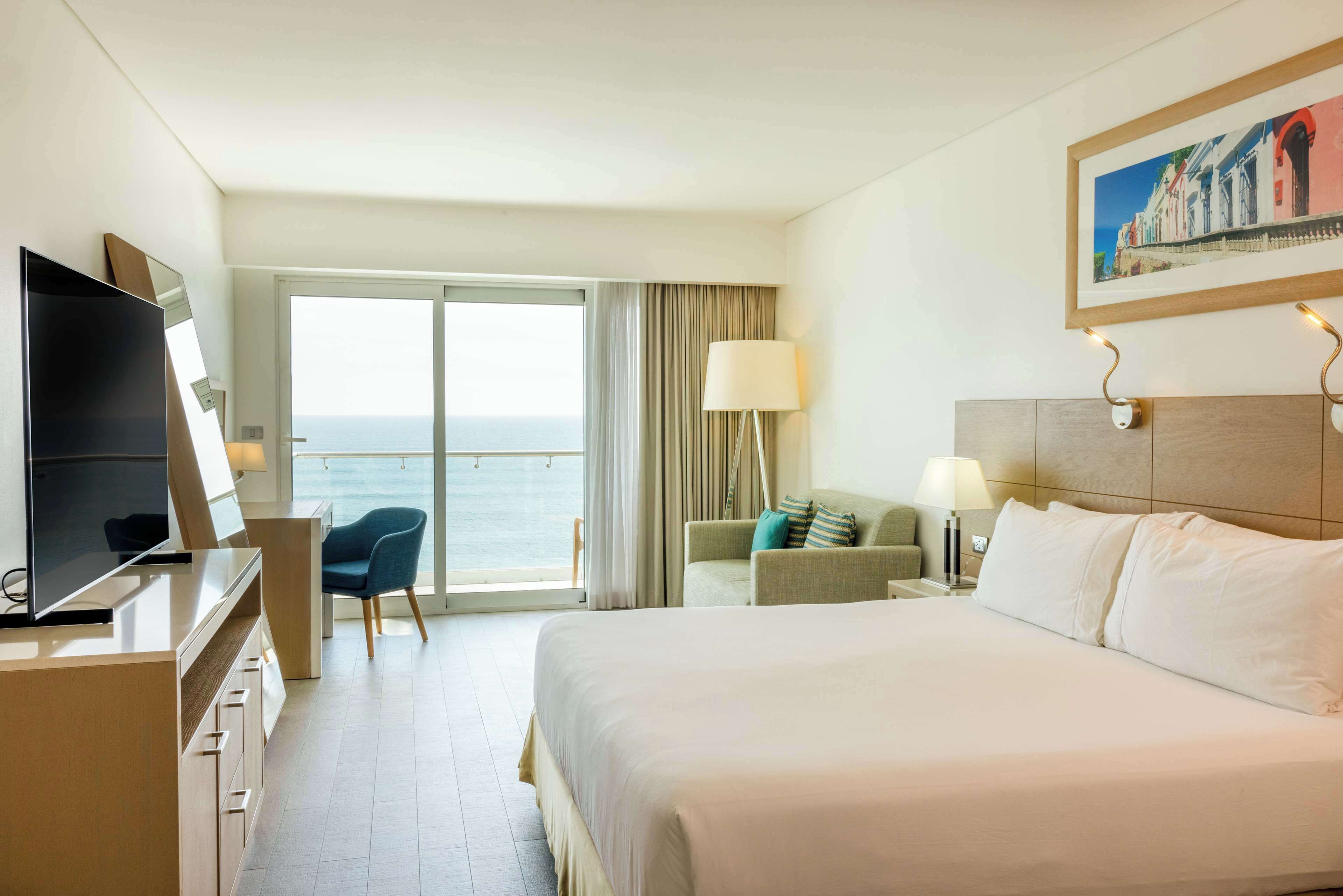 A hotel room with a large bed, a desk with a TV, a chair, a sofa, a lamp, and a painting on the wall.