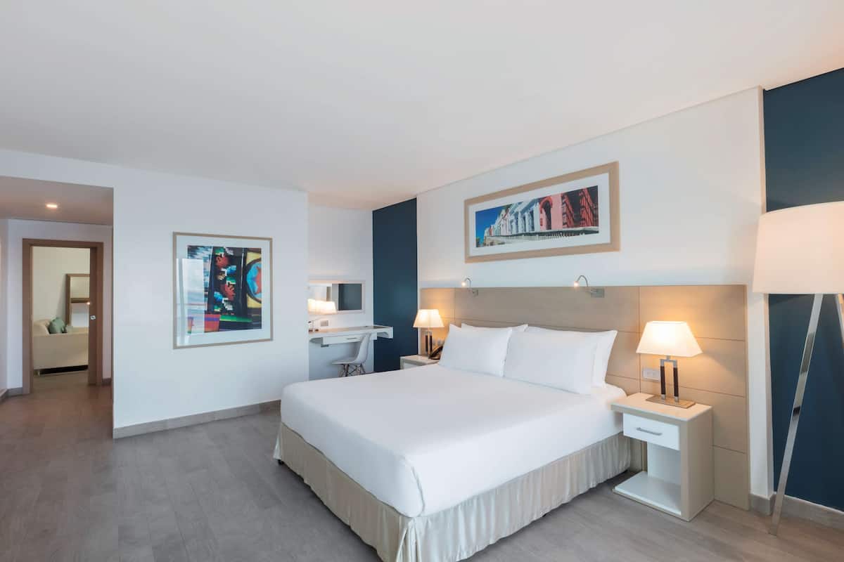 Habitación de hotel con una cama grande, mesitas de noche con lámparas, un escritorio con una silla y obras de arte en las paredes.