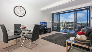 A modern living room with a glass dining table, black chairs, a flat-screen TV, and a large window offering a city view. - Accommodate Canberra - Dockside (Kingston)