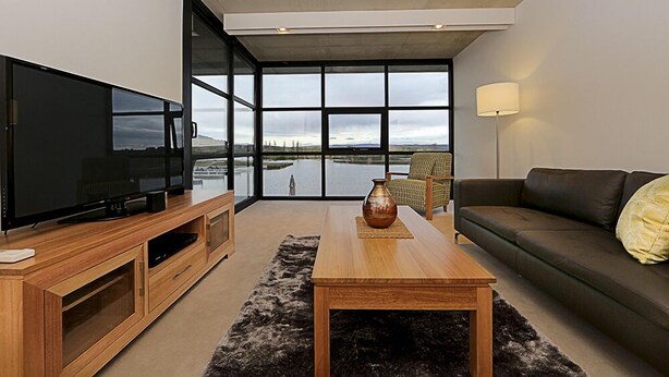 A modern living room with a large flat-screen TV, wooden furniture, a coffee table, and a view of a lake through large windows. - Accommodate Canberra - Dockside (Kingston)