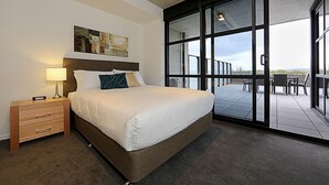 A modern hotel room with a large bed, a nightstand, a lamp, and a view through large glass doors to an outdoor patio area. - Accommodate Canberra - Dockside (Kingston)