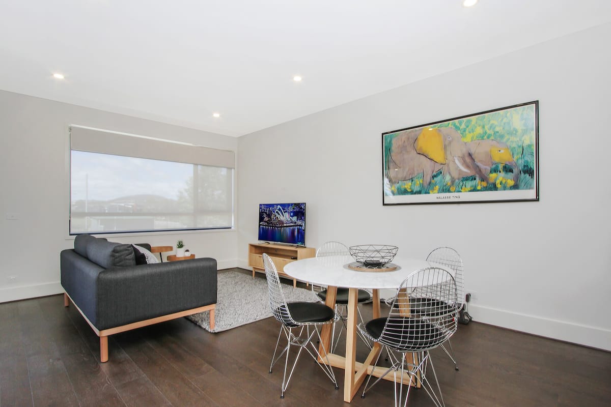 A modern living room with a gray sofa, a round dining table with wire chairs, a large window, and a framed painting on the wall.
