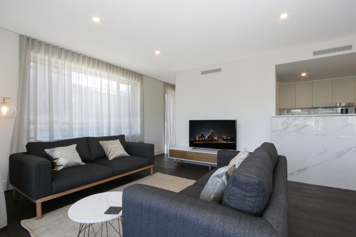 A modern living room with a dark grey sofa, a white coffee table, and a flat-screen TV on a wooden stand.