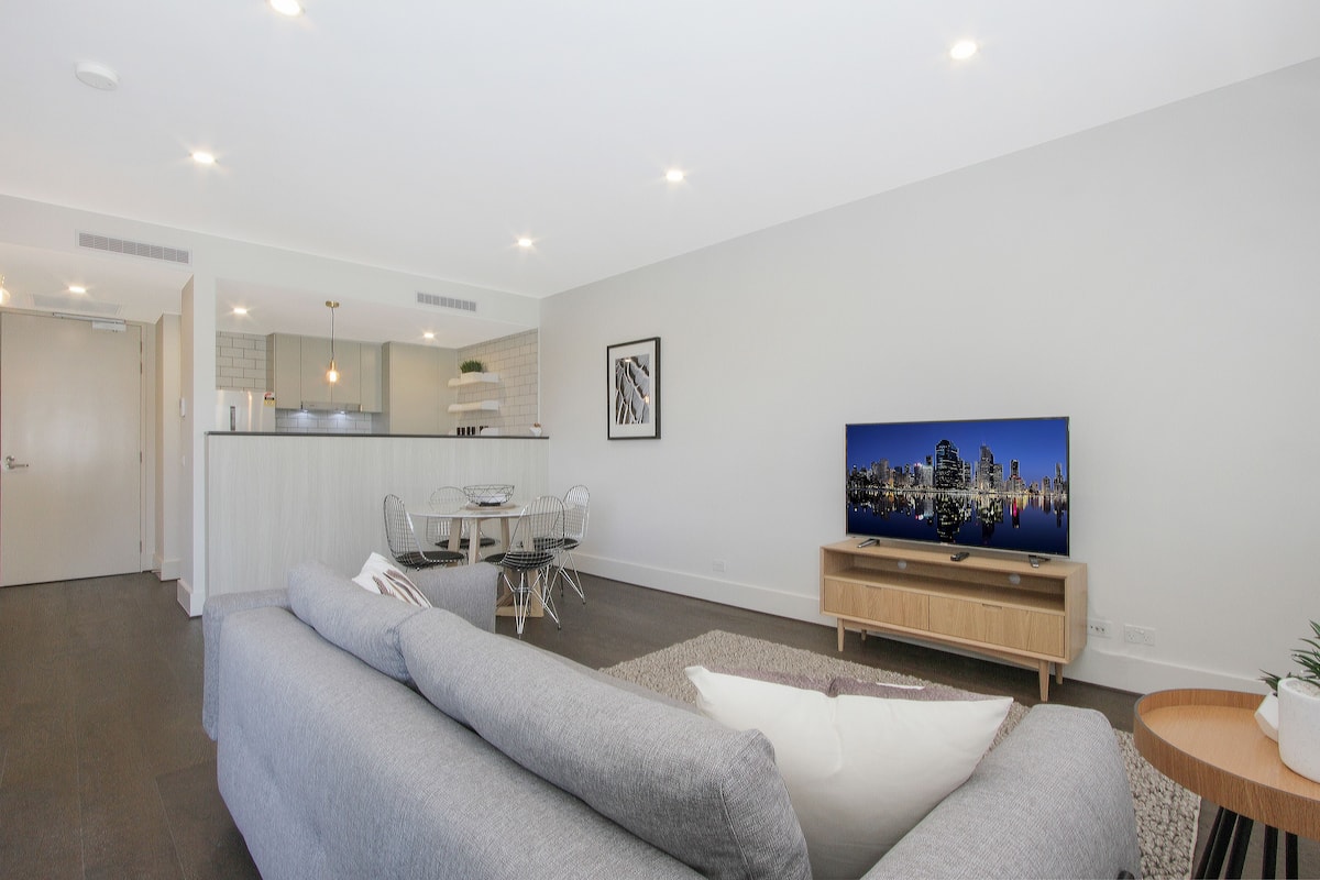 A modern living room with a grey sofa, a TV, and a dining area with chairs and a table.