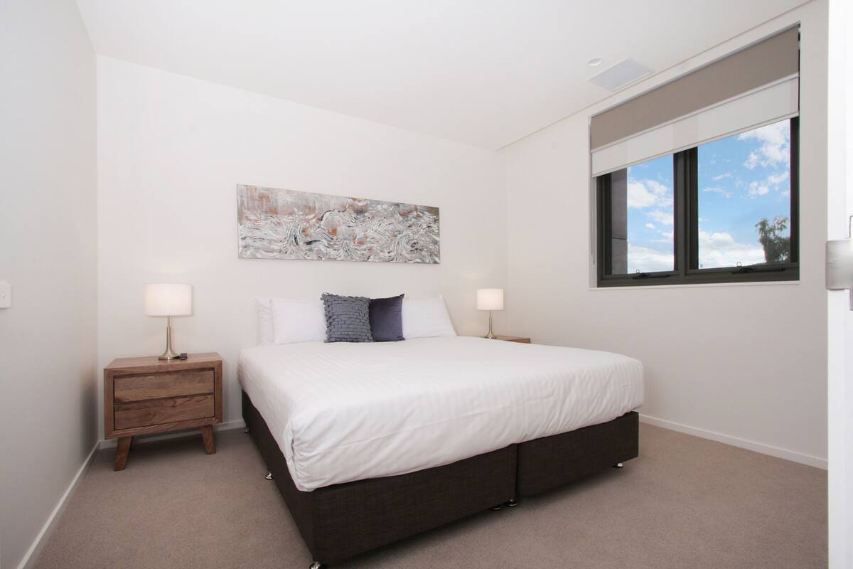 A bedroom with a large bed, two bedside tables, a window with blinds, and a wall-mounted abstract painting.