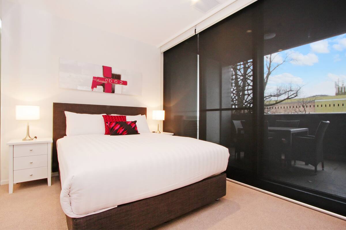 A modern hotel room with a large bed, two bedside tables with lamps, a window with a view of the outdoors, and a wall-mounted artwork.