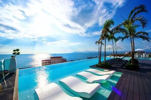 Ein Infinity-Pool mit Blick auf den Ozean, Palmen und eine Holzterrasse mit Liegestühlen.