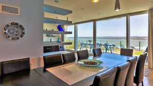A modern dining area with a large table and chairs, a decorative plate, and a view of the sea through large windows.