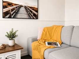 Una sala de estar con un sofá gris, una manta amarilla, una revista y un cuadro con una imagen de un puente de madera.