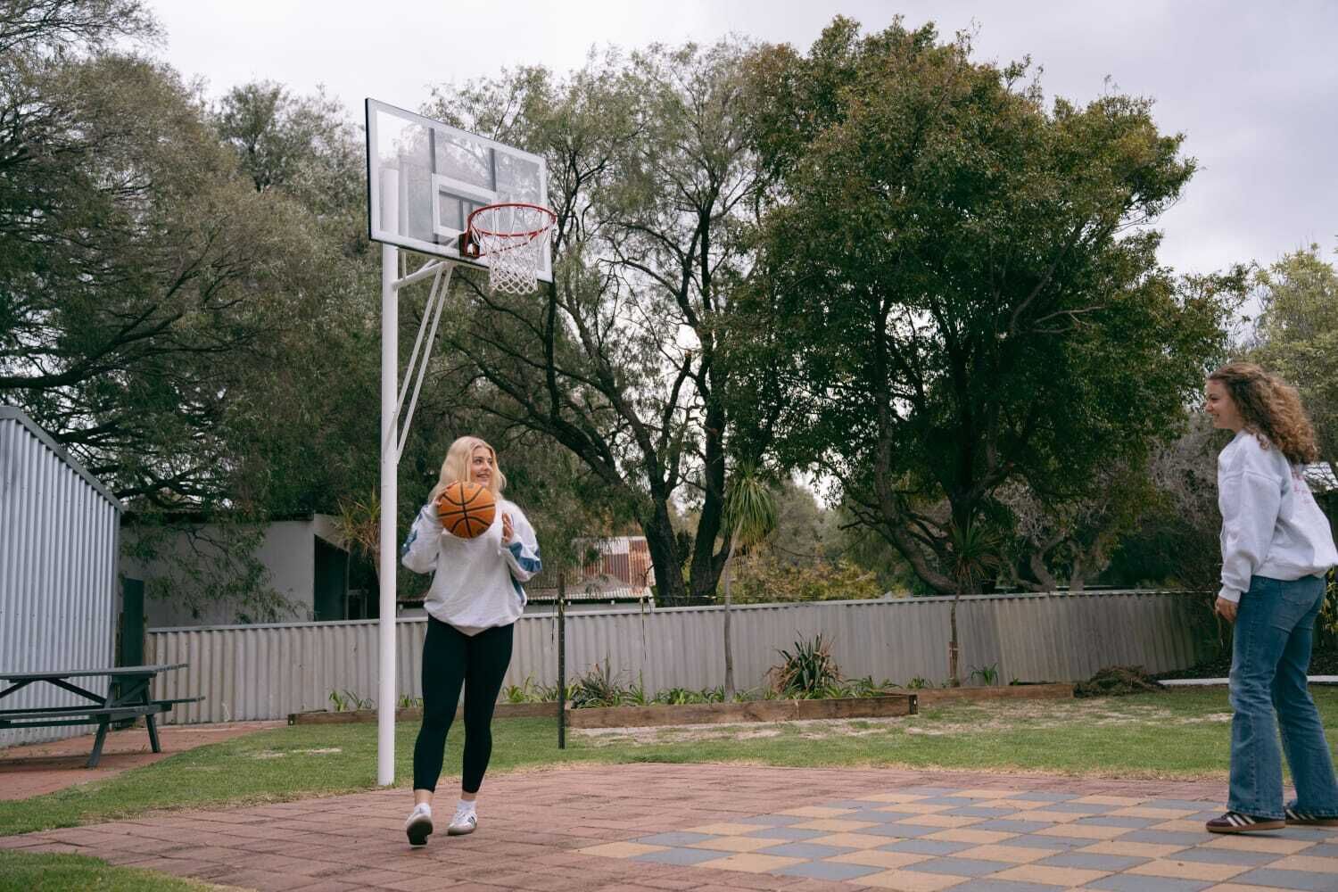 Un terrain de basket en plein air, avec un panier, une table de pique-nique et une zone délimitée par une clôture, agrémentée d’arbres.