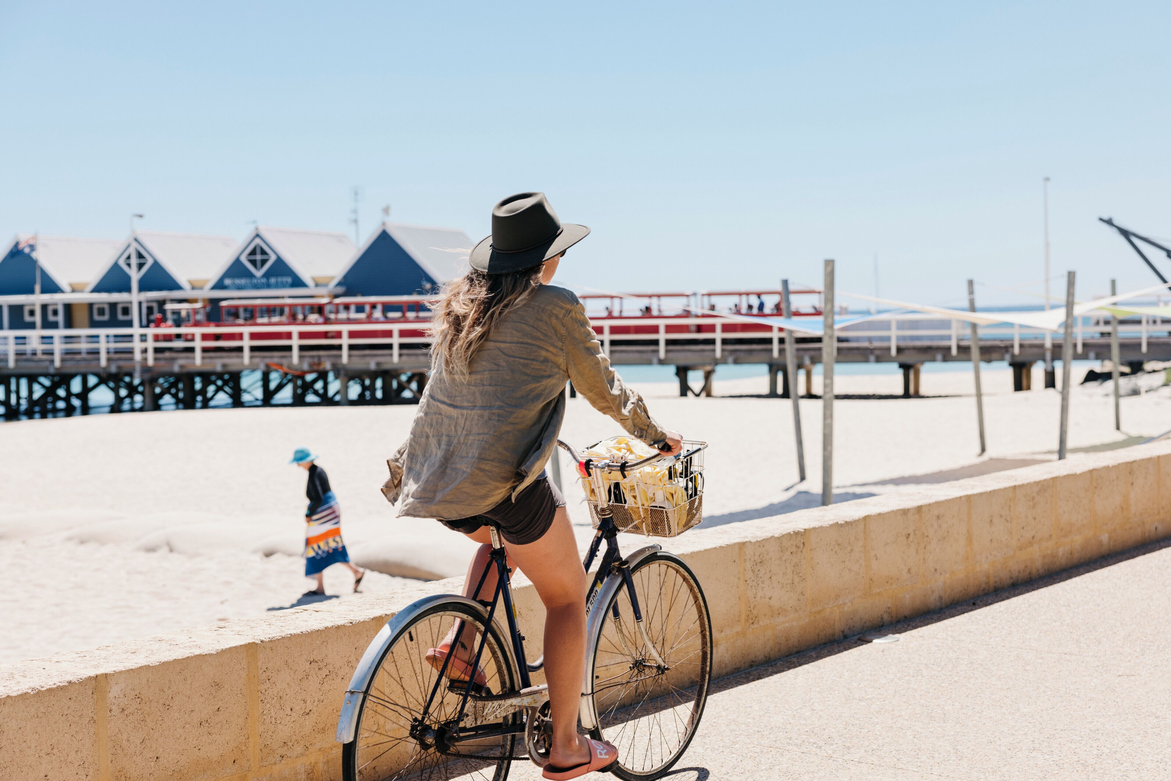 Una persona pedaleando en bicicleta con una cesta por un sendero arenoso cerca de un muelle.