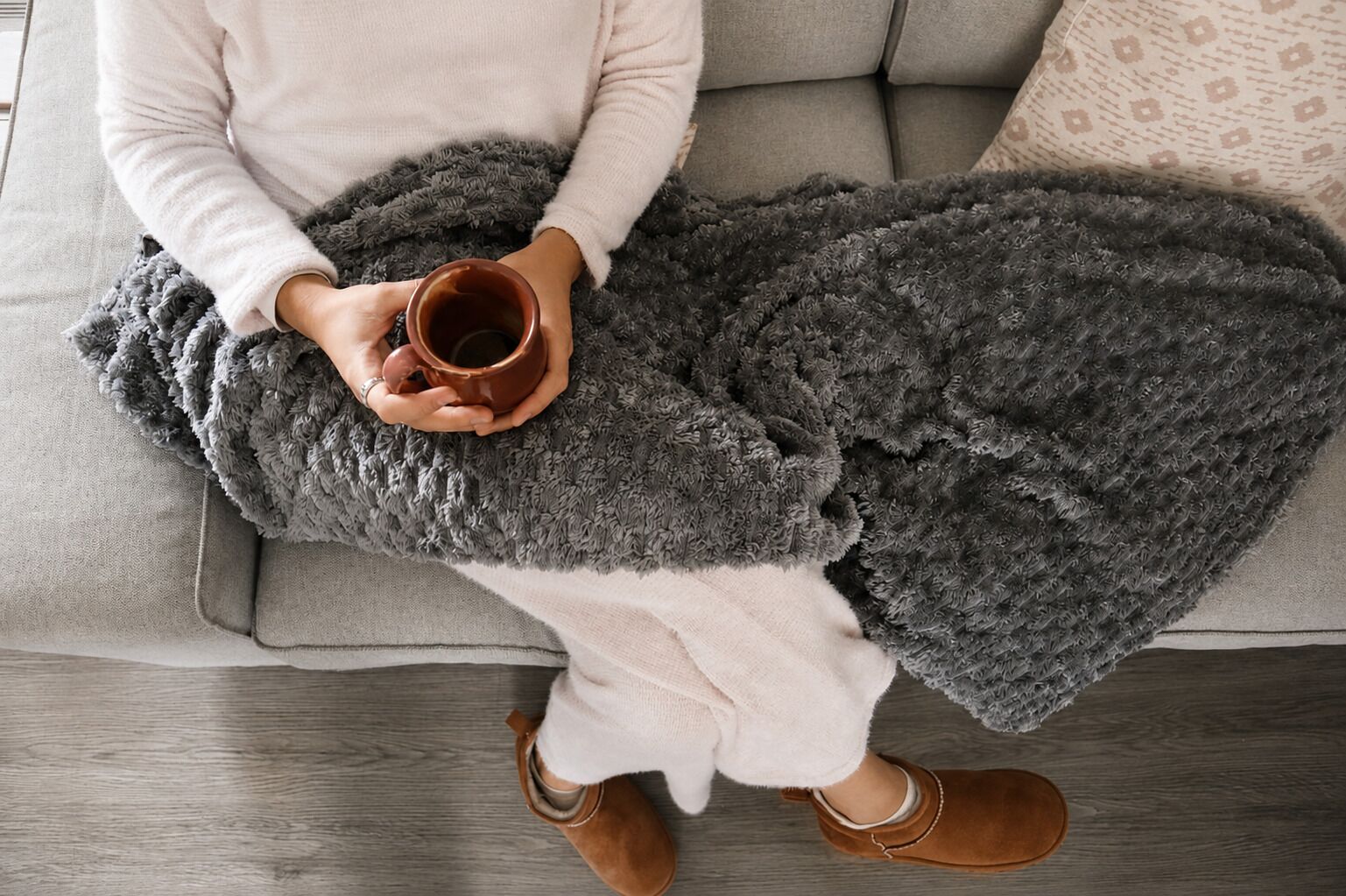 Eine Person sitzt auf einem grauen Sofa mit einer grauen Decke und hält eine braune Tasse.