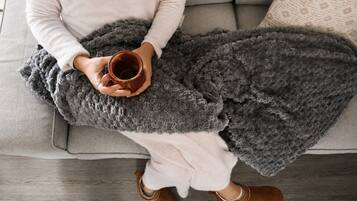 Eine Person sitzt auf einem grauen Sofa mit einer grauen Decke und hält eine braune Tasse.