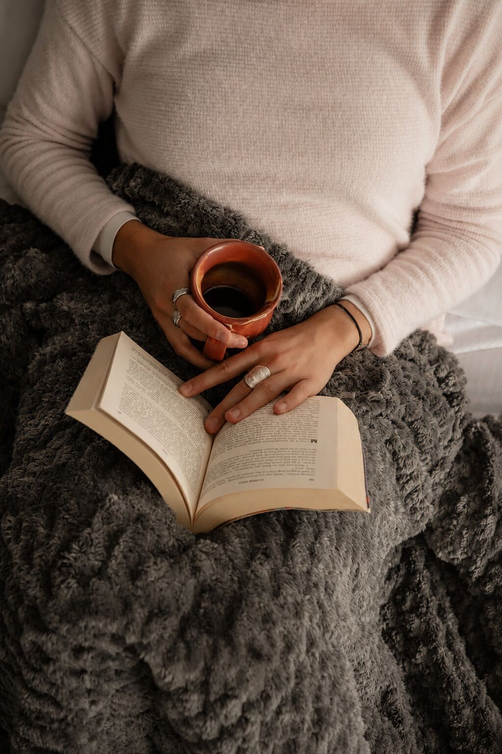 A person sitting with a book and a cup, on a grey blanket.