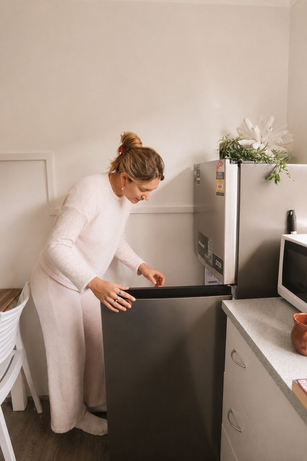 A person opening a refrigerator door in a kitchen with a microwave and a small potted plant on the counter.