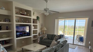 A living room with a grey sofa, a flat-screen TV, shelves with decorative items, and a view of a swimming pool through large glass doors. - Villa With Private Pool And Jacuzzi, Ideally Situated for Disney (Clermont)