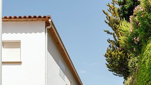 Un chemin mène à un bâtiment blanc surmonté d’un toit en tuiles, entouré de haies soigneusement taillées et d’une végétation luxuriante.