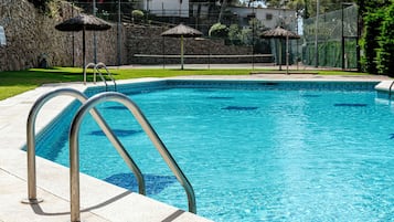 Une piscine aux eaux cristallines, dotée d’échelles en métal, un mur en pierre et une zone herbeuse agrémentée de parasols.