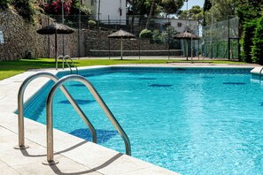 A clear swimming pool with metal ladders, a stone wall, and a grassy area with umbrellas.