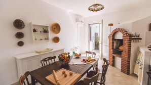 A dining area with a wooden table, chairs, and a variety of fruits and vegetables. There is a brick oven in the background and a white cabinet with decorative plates on the wall. - Villa Ferradina 4+2, Emma Villas (Ispica (Ragusa))