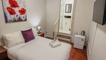 A hotel room with a bed, a nightstand, a lamp, a painting, a mirror, a shower, and a small refrigerator.