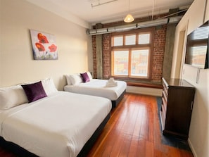A hotel room with two beds, a brick wall, a window with blinds, and a wooden dresser. - Aura Hotel (Wellington)