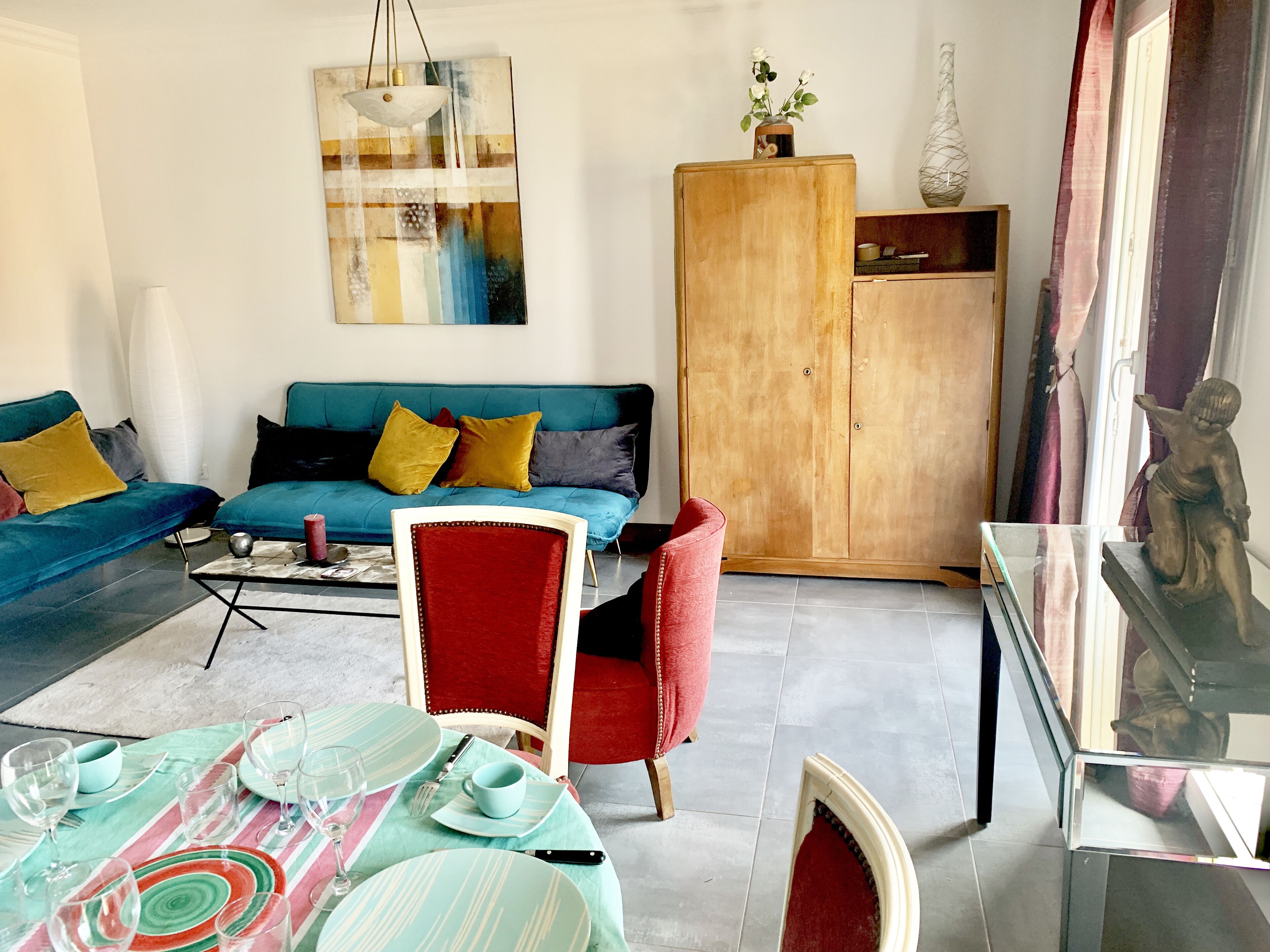 A living room with a blue sofa, red chair, dining table set with plates and glasses, and a wooden cabinet.