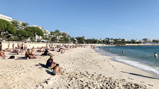 A crowded beach with people sunbathing and swimming, palm trees, and buildings in the background.