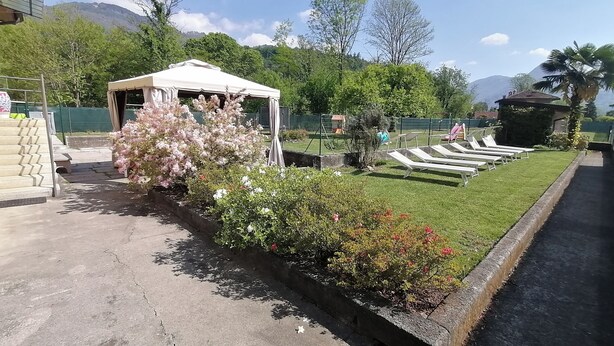 Un giardino con una tenda bianca, un vialetto e un'area piscina con poltrone relax.