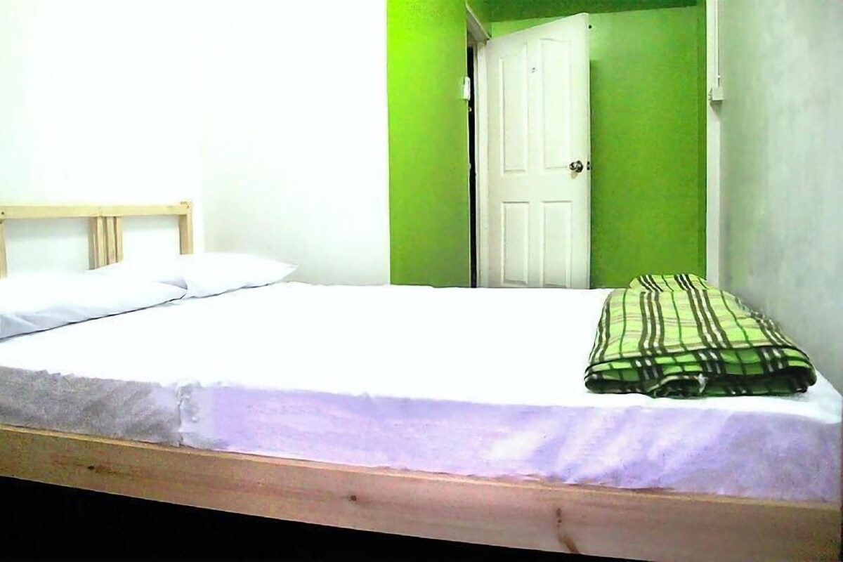 Un lit simple avec une tête de lit en bois, un matelas blanc et une couverture à carreaux verts et blancs.