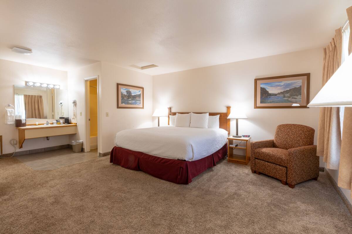 A hotel room with a large bed, a chair, a vanity with a mirror, and two framed pictures on the wall.
